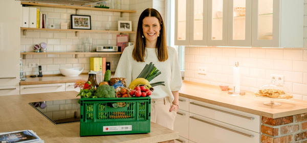KI generiert: Eine Frau steht in einer Küche neben einer Kiste voller Obst und Gemüse.