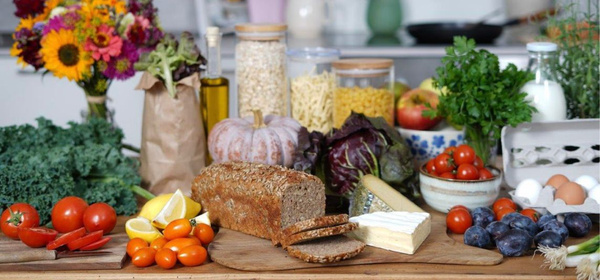 KI generiert: Verschiedene frische Lebensmittel auf einem Tisch: Brot, Gemüse, Obst, Eier und Blumenstrauß.